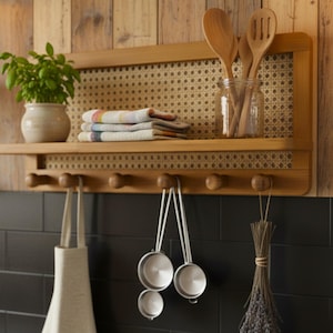 Puede incluir: Estante de cocina de madera con respaldo tejido, que contiene una planta en maceta, toallas dobladas y un tarro de cristal con cucharas de madera. Debajo, ganchos sostienen un delantal, tazas medidoras y un manojo de hierbas secas.