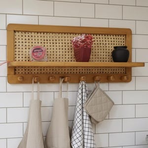 Puede incluir: Estante de cocina de madera con respaldo de mimbre y ganchos. Exhibe una maceta roja con flores, un jarr&oacute;n negro y un rollo de cinta adhesiva. Delantales, un pa&ntilde;o de cocina a cuadros y un agarrador cuelgan debajo.