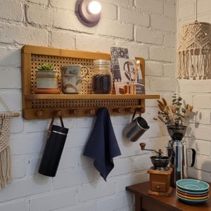 Puede incluir: Una estanter&iacute;a de pared de madera con respaldo de ca&ntilde;a tejida, que contiene una peque&ntilde;a planta en maceta, tazas y un tarro de frijoles. Debajo, ganchos sostienen un termo negro, un pa&ntilde;o azul y una jarra de metal. Un molinillo de caf&eacute; y platos apilados est&aacute;n en una mesa cercana.