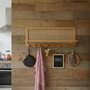 Puede incluir: Un organizador de pared de cocina de madera con un panel de mimbre tejido, un estante y ganchos. Una sart&eacute;n de hierro fundido negra, una toalla a cuadros rojos y blancos, una peque&ntilde;a pizarra con "COFFEE" y una tabla de cortar de madera cuelgan de los ganchos.