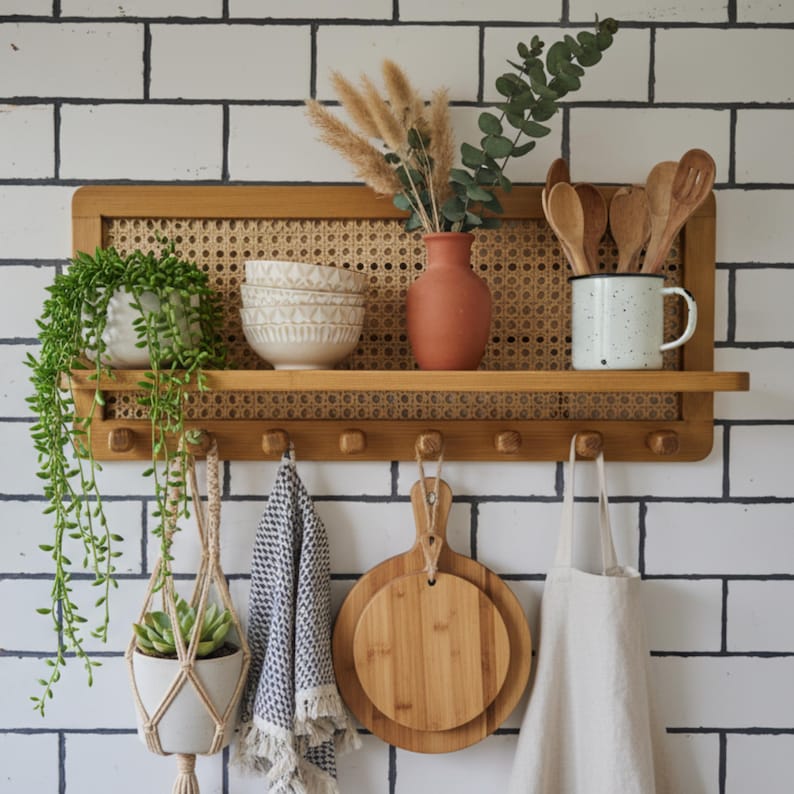 Puede incluir: Una estanter&iacute;a de cocina de madera con respaldo tejido, que contiene cuencos decorativos, un jarr&oacute;n de terracota y una taza moteada. Debajo, una planta colgante, una toalla estampada, tablas de cortar y un delantal cuelgan de ganchos.
