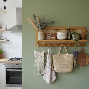 Puede incluir: Una estanter&iacute;a de madera con respaldo de ca&ntilde;a tejida est&aacute; montada en una pared verde. La estanter&iacute;a contiene cuencos, una botella de aceite de oliva y una planta peque&ntilde;a. Debajo, un tapiz de macram&eacute;, un delantal floral, un bolso de mano de paja y una tabla de cortar de madera.