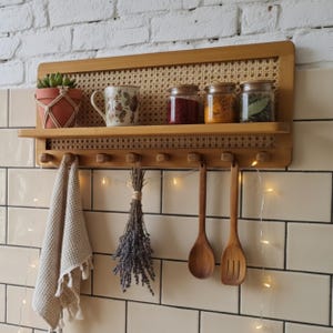 Puede incluir: Estante de cocina de madera con respaldo de mimbre, que contiene una planta en maceta, una taza floral y frascos de especias. Debajo, una toalla, lavanda y cucharas de madera cuelgan de clavijas.