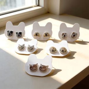 May include: White cat-shaped earring holders with pairs of dog and cat stud earrings. The earrings feature realistic images of dogs and cats. The earrings are in various colors, including brown, black, and gray.