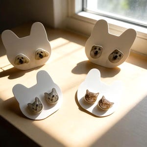 May include: Four pairs of earrings are displayed on a wooden surface. The earrings are white, cat-shaped cards with dog and cat face studs. The dog earrings have golden retriever faces, and the cat earrings have tabby cat faces.