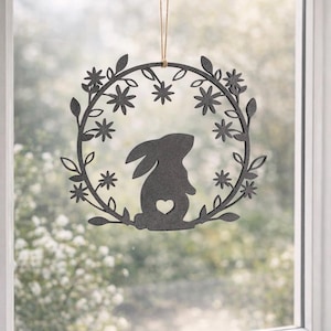 Könnte beinhalten: Dunkelgraue Ostersilhouette eines Hasen mit Herz-Ausschnitt, zentriert in einem floralen Kranz. Der Kranz ist mit Blättern und sternförmigen Blüten verziert, aufgehängt an einem natürlichen Bindfaden. Das Design wird vor einem verschwommenen Außenhintergrund gezeigt.