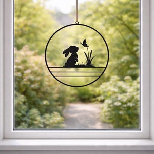 Könnte beinhalten: Schwarze Metall-Wandkunst in Kreisform mit einer Silhouette eines Kaninchens, das einen Schmetterling betrachtet. Das Design umfasst Gras und eine horizontale Linie am unteren Rand. Die Kunst wird von einer dünnen, braunen Schnur gehalten, vor einem verschwommenen Hintergrund.