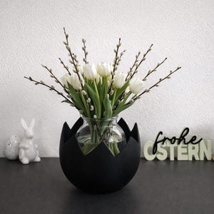 Könnte beinhalten: Eine schwarze, eiförmige Vase hält weiße Tulpen und Weidenkätzchenzweige. Eine Glasvase befindet sich in der schwarzen Vase. Ein weißer Keramik-Hase und ein Ei befinden sich links. Eine brennende Kerze und ein Schild mit der Aufschrift "frohe OSTERN" befinden sich rechts.