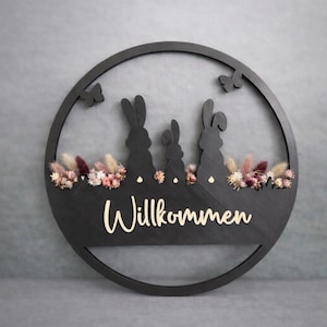 Könnte beinhalten: Schwarzes, rundes Wandbild aus Holz mit einer Hasenfamilien-Silhouette und dem Wort "Willkommen" in Schreibschrift. Das Design umfasst Trockenblumen und Schmetterlingsausschnitte und schafft eine rustikale Dekoration zum Thema Frühling.