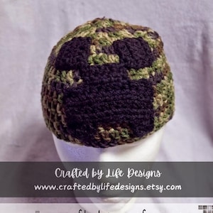 May include: A crocheted hat in a camouflage pattern with a black crocheted design. The hat is on a white mannequin head. The text "Crafted by Life Designs" is on a black banner below the hat. The text "www.craftedbylifedesigns.etsy.com" is on the banner below the first text. The text "Crafted by Life Designs" is on a black banner below the website address.