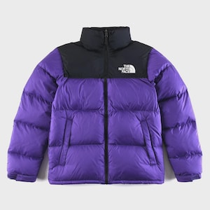 Könnte beinhalten: Gesteppte Winterjacke in Lila und Schwarz. Die Jacke hat einen hohen Kragen und einen Reißverschluss. Das The North Face-Logo befindet sich auf der linken Brust. Für kaltes Wetter und Outdoor-Aktivitäten konzipiert.