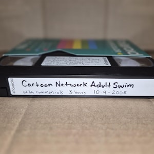 May include: A black Polaroid VHS tape with a white label on the front edge featuring handwritten text that reads: Cartoon Network Adult Swim, With Commercials, 3 hours, 10-9-2005. The tape is partially inserted into a teal and white cardboard sleeve.