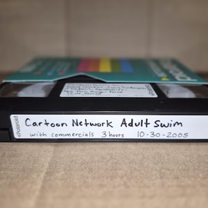 May include: A black VHS tape with a white label on the front edge featuring the handwritten text Cartoon Network Adult Swim with commercials 3 hours 10-30-2005. A second white label on the top surface contains additional handwritten program details.