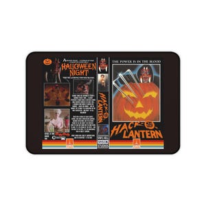 May include: A rectangular mat featuring a vintage VHS cover design for the horror film Halloween Night and Hack-O-Lantern. The black background displays orange text, movie stills, a carved pumpkin with sharp claws, and the Vinyl Hell Studios logo.