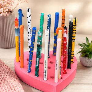 Peut inclure: Un porte-stylo rose en forme de cœur avec de multiples trous, contenant une collection de stylos colorés assortis avec divers motifs de personnages, des imprimés et du texte incluant "Rise and Shine", "Rock 'n Rolling" et "Be Curious".
