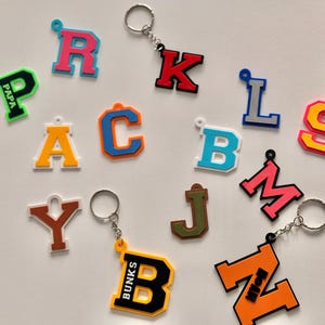 Peut inclure: Une collection de porte-clés lettres colorés, chacun avec un porte-clés argenté. Les lettres sont de différentes couleurs, notamment rose, bleu, orange, jaune et vert. Certains porte-clés comportent du texte supplémentaire, comme "PAPA" et "BUNKS".