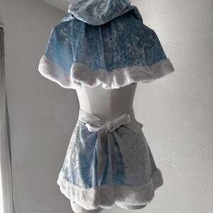 Puede incluir: Un conjunto de disfraz de terciopelo azul claro con una capa con capucha y una falda a juego. Los bordes de la capa y la falda est&aacute;n adornados con piel sint&eacute;tica blanca. Una cinta de sat&eacute;n blanco se ata a la cintura. El disfraz se exhibe en un maniqu&iacute;.