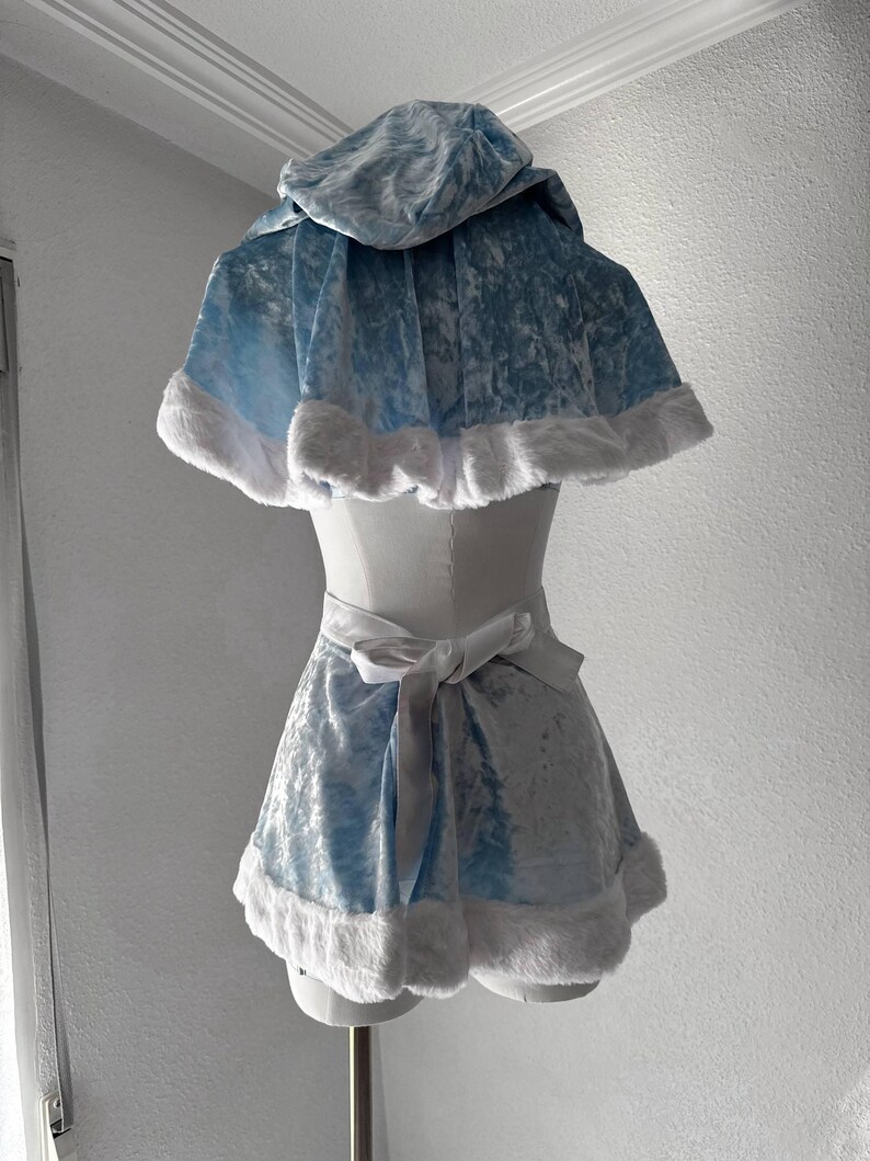 Puede incluir: Un conjunto de disfraz de terciopelo azul claro con una capa con capucha y una falda a juego. Los bordes de la capa y la falda est&aacute;n adornados con piel sint&eacute;tica blanca. Una cinta de sat&eacute;n blanco se ata a la cintura. El disfraz se exhibe en un maniqu&iacute;.