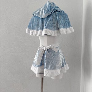 Puede incluir: Un conjunto de disfraz de terciopelo azul claro con una capa con capucha y pantalones cortos a juego, con ribete de piel sint&eacute;tica blanca. Los pantalones cortos tienen un lazo blanco en la cintura. El atuendo se muestra en un maniqu&iacute;.