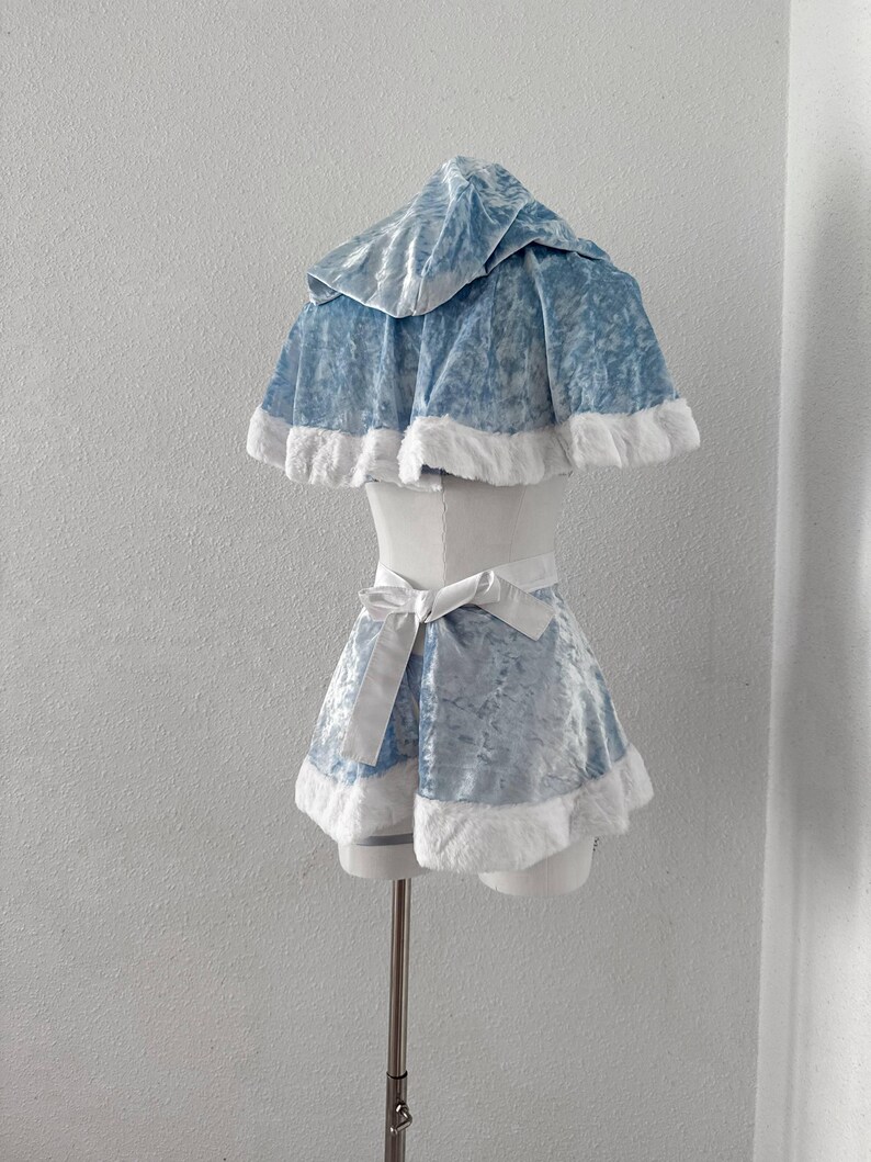 Puede incluir: Un conjunto de disfraz de terciopelo azul claro con una capa con capucha y pantalones cortos a juego, con ribete de piel sint&eacute;tica blanca. Los pantalones cortos tienen un lazo blanco en la cintura. El atuendo se muestra en un maniqu&iacute;.
