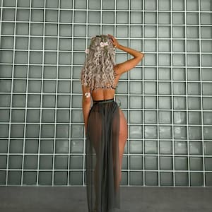 May include: A woman's back is shown, wearing a black top and a sheer, olive-green skirt. Her long, curly, silver hair is adorned with pink flowers. She is standing in front of a gray, grid-patterned wall.
