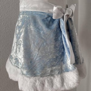 Puede incluir: Una falda de terciopelo azul claro con una cinturilla y lazo de sat&eacute;n blanco. El dobladillo de la falda est&aacute; adornado con suave piel sint&eacute;tica blanca. La prenda se exhibe en un maniqu&iacute;, mostrando su dise&ntilde;o y textura.