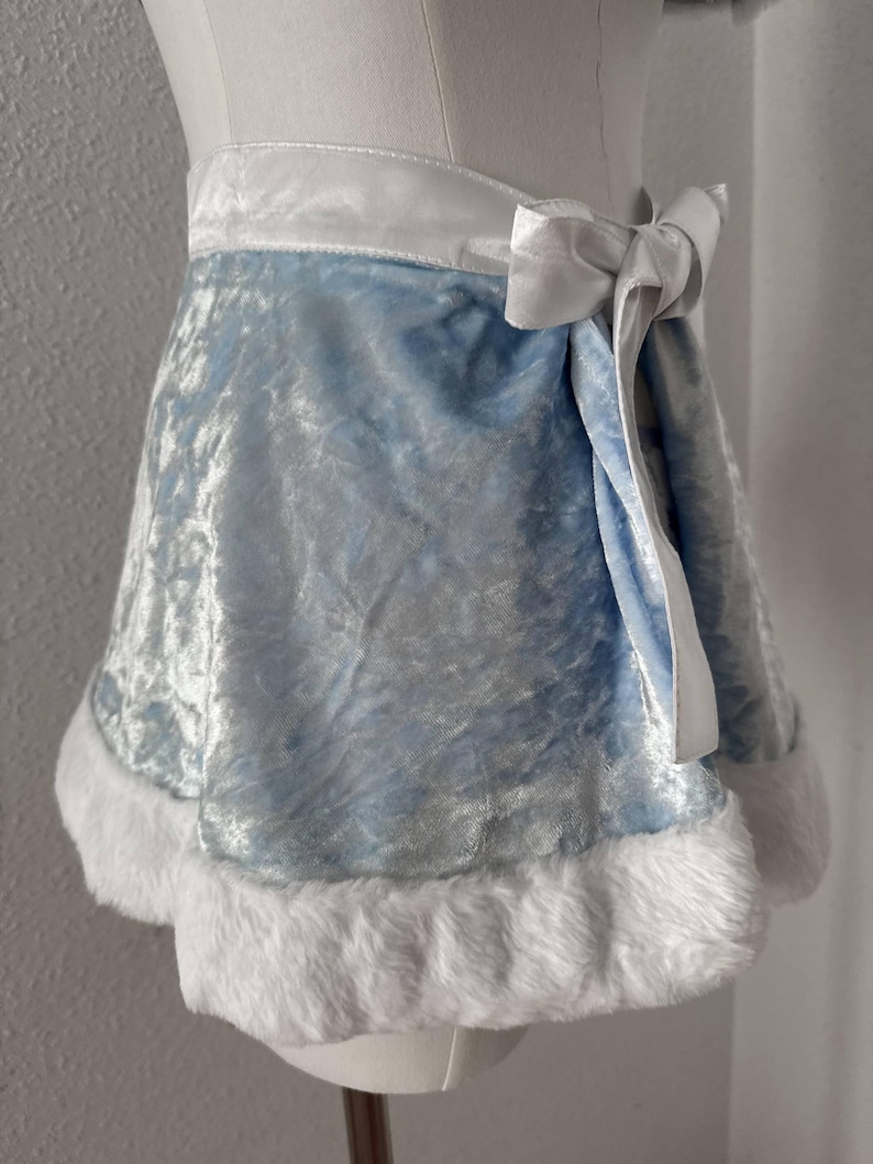 Puede incluir: Una falda de terciopelo azul claro con una cinturilla y lazo de sat&eacute;n blanco. El dobladillo de la falda est&aacute; adornado con suave piel sint&eacute;tica blanca. La prenda se exhibe en un maniqu&iacute;, mostrando su dise&ntilde;o y textura.