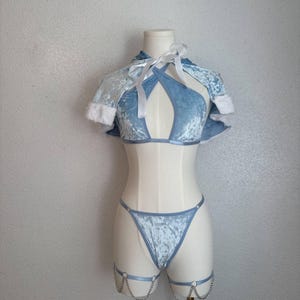 Puede incluir: Conjunto de lencer&iacute;a de terciopelo azul claro sobre un maniqu&iacute;. El conjunto incluye un top halter con capa, un tanga y ligas con detalles de cadena. La capa tiene ribete de piel sint&eacute;tica blanca y lazos blancos.