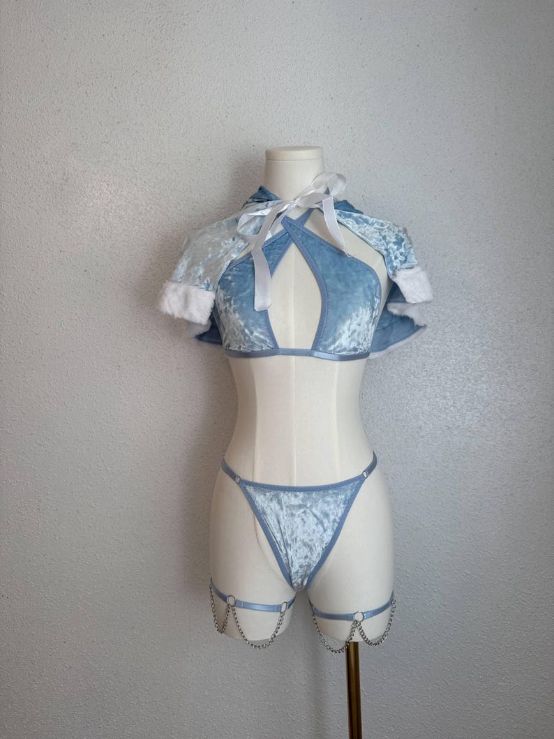Puede incluir: Conjunto de lencer&iacute;a de terciopelo azul claro sobre un maniqu&iacute;. El conjunto incluye un top halter con capa, un tanga y ligas con detalles de cadena. La capa tiene ribete de piel sint&eacute;tica blanca y lazos blancos.