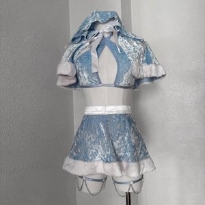 Puede incluir: Un conjunto de disfraz de terciopelo azul claro con ribete de piel sint&eacute;tica blanca. El conjunto incluye una capa con capucha, un top corto y una minifalda. La falda presenta ligas adjuntas con detalles de cadena. El atuendo est&aacute; dise&ntilde;ado para un evento festivo o tem&aacute;tico.