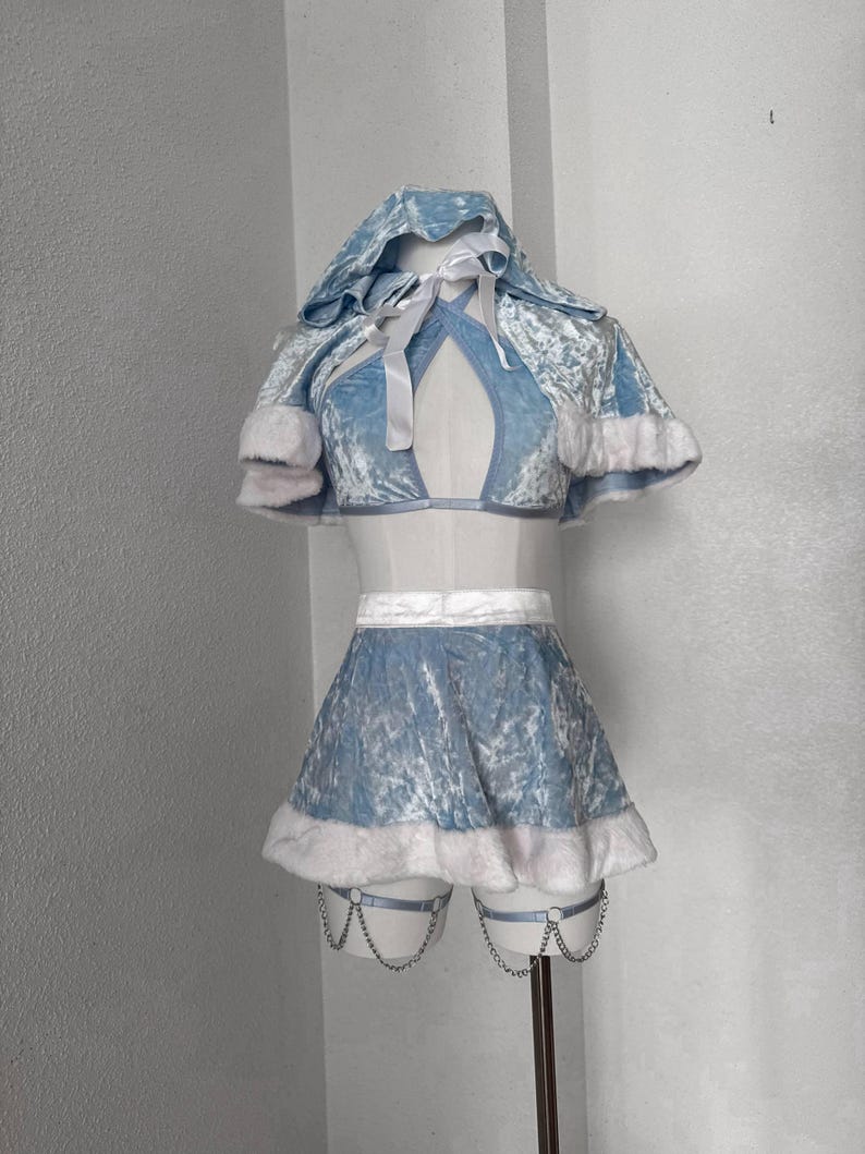 Puede incluir: Un conjunto de disfraz de terciopelo azul claro con ribete de piel sint&eacute;tica blanca. El conjunto incluye una capa con capucha, un top corto y una minifalda. La falda presenta ligas adjuntas con detalles de cadena. El atuendo est&aacute; dise&ntilde;ado para un evento festivo o tem&aacute;tico.