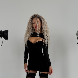 Op de afbeelding: Een zwarte mini-jurk met lange mouwen en een uitgesneden detail bij de borst. De jurk heeft een sweetheart-halslijn en wordt gecombineerd met zwarte dijhoge kousen met jarretels. Het model heeft lang, krullend blond haar.