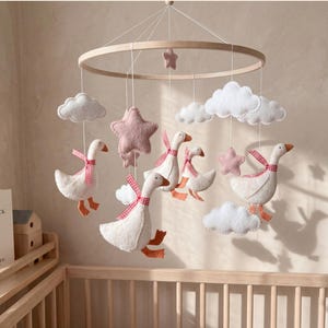 Pode incluir: Móvel de bebê artesanal com gansos brancos, estrelas rosa e nuvens brancas. Os gansos têm laços xadrez vermelho e branco. O móvel está pendurado em um anel de madeira acima de um berço. Um livro infantil é visível.