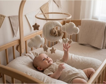 Könnte beinhalten: Ein Baby in einem Holzbettchen mit einem beigen Mobile mit Wolken, Sternen und einem Mond mit einem Teddybären. Das Bettchen hat eine beige Matratze und eine Decke. Ein Banner aus beigen Dreiecken hängt an der Wand.