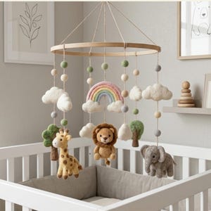 Op de afbeelding: Een baby mobile met een houten ring en hangende vilten dierenfiguren, waaronder een giraf, leeuw en olifant. De mobile heeft ook vilten wolken, een regenboog en kleurrijke vilten ballen. Het hangt boven een wit babybedje.