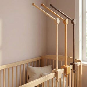 Puede incluir: Brazo de móvil de cuna de madera en varios tonos de marrón. El brazo del móvil se fija a la barandilla de la cuna con una abrazadera. El brazo del móvil tiene una varilla larga y delgada de madera con una pieza circular de madera en el extremo.