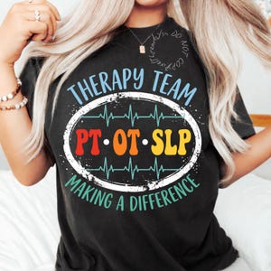 May include: A black t-shirt featuring a graphic with the text Therapy Team in light blue, PT OT SLP in red, orange, and yellow, and Making A Difference in teal. The design includes a white oval border and a teal heartbeat line across the centre.