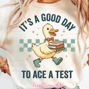 May include: Cream-colored t-shirt with a cartoon duck carrying books, wearing a bow tie and boots. Text reads "IT'S A GOOD DAY TO ACE A TEST" in teal. The design includes stars and a checkered pattern.