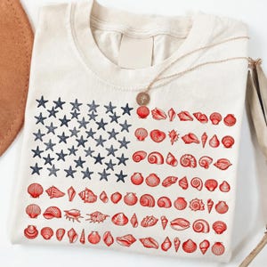 May include: A cream-colored t-shirt featuring a graphic design arranged like the American flag. The canton consists of dark blue starfish, while the stripes are composed of various red seashells. A gold-toned necklace rests on the neckline.