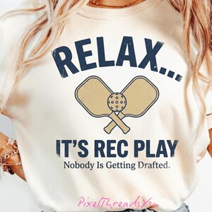 Könnte beinhalten: Cremefarbenes T-Shirt mit dem Schriftzug "RELAX..." über einer Grafik mit gekreuzten Pickleball-Schlägern und einem Ball. Darunter steht der Text "IT'S REC PLAY" und "Nobody Is Getting Drafted."
