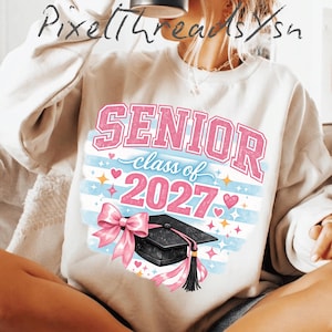 May include: A cream-colored crewneck sweatshirt featuring a graphic print with the text SENIOR class of 2027 in pink and blue. The design includes a black graduation cap with a pink bow, surrounded by small pink hearts and yellow star accents.