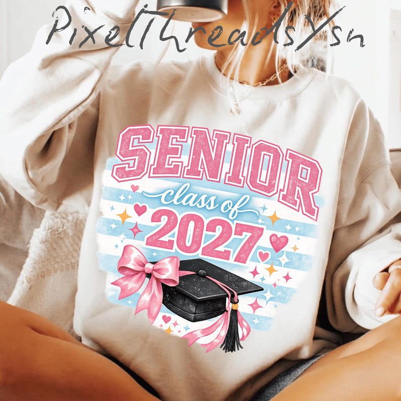May include: A cream-colored crewneck sweatshirt featuring a graphic print with the text SENIOR class of 2027 in pink and blue. The design includes a black graduation cap with a pink bow, surrounded by small pink hearts and yellow star accents.