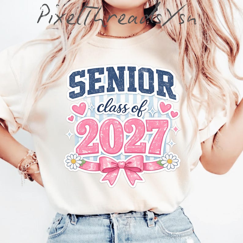 May include: A cream-colored t-shirt featuring a graphic with the text "SENIOR class of 2027" in dark blue and pink. The design includes pink hearts, a large pink bow, and white daisies against a light blue striped background with star accents.