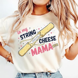 May include: Cream-colored sweatshirt with a graphic of a string cheese, partially pulled apart. Text reads "in my STRING CHEESE MAMA era" in pink and black. Small stars and hearts are scattered around the design.