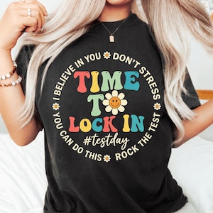 May include: Black t-shirt with a colorful graphic. The design includes the words "I BELIEVE IN YOU" and "DON'T STRESS" around the top. The words "TIME TO LOCK IN" are in the center, with a daisy and the hashtag "#testday".