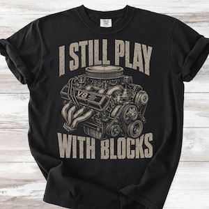 Könnte beinhalten: Ein schwarzes T-Shirt mit kurzen Ärmeln und einer beigen Grafik eines V8-Motors. Der Text "I STILL PLAY WITH BLOCKS" ist in einer abgenutzten beigen Schrift über und unter der Motorabbildung gedruckt. Das Shirt hat einen Rundhalsausschnitt.