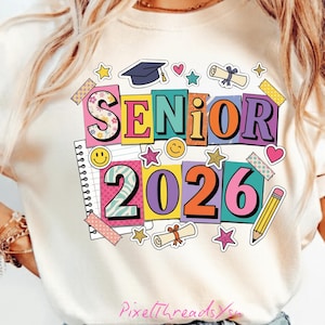 Op de afbeelding: Crèmekleurig T-shirt met een kleurrijke "SENIOR 2026" afbeelding. Het ontwerp bevat een afstudeerhoed, sterren, harten, een diploma, een potlood en smiley gezichten. De tekst is in verschillende heldere kleuren en lettertypen.