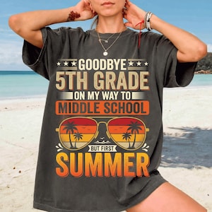Op de afbeelding: Donkergrijs t-shirt met de tekst "Goodbye 5th Grade On My Way To Middle School But First Summer". Het ontwerp bevat een zonnebril met palmbomen en een zonsondergang. Het shirt heeft een casual stijl.