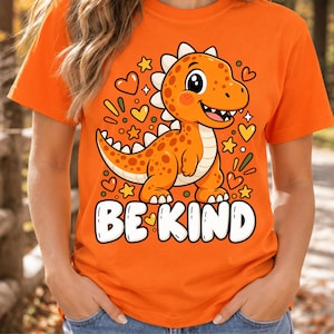 Op de afbeelding: Een oranje t-shirt met een afbeelding van een lachende oranje dinosaurus met witte stekels, omringd door gele sterren en oranje hartjes. Onder de dinosaurus staat de tekst BE KIND in grote, dikke, witte hoofdletters.