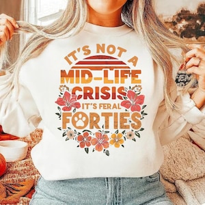 Op de afbeelding: Witte sweatshirt met de tekst "It's Not a Mid-Life Crisis, It's Feral Forties" in oranje en geel. Het ontwerp bevat een zonsondergang, bloemen en een voetafdruk.